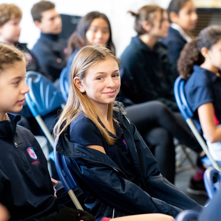 A smiling student sat on the chair, with other students sat on their chairs, as they sit ready for a group discussion or assembly