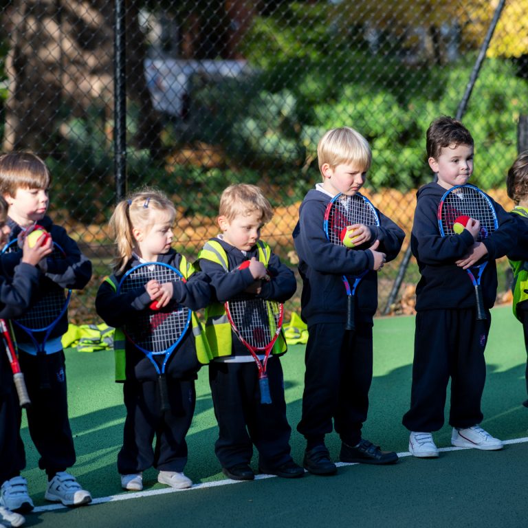 5 children stood in a row holding tennis rackets and balls in their hands