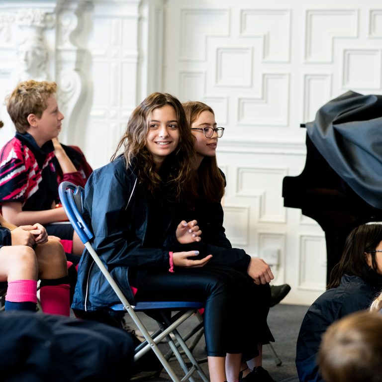 A student sat on the table looking over, surrounded by other students on chairs and sat on the floor